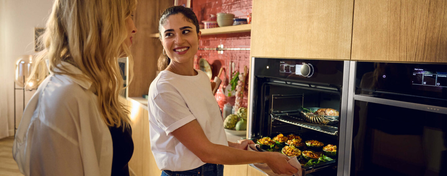 Zwei Frauen stehen in moderner Küche von Neff, während eine ein Blech mit gefülltem Gemüse aus dem Backofen nimmt und beide sich lächelnd unterhalten bei A2 Küchen